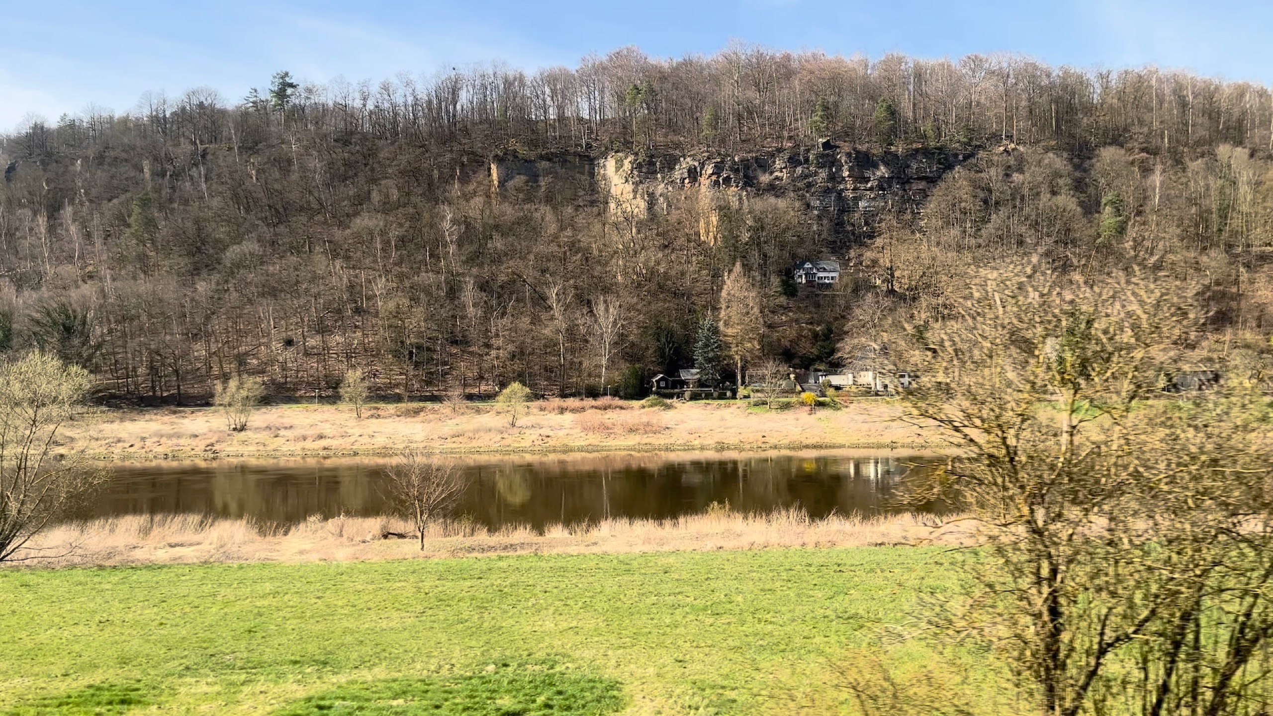 Blick aus dem Bahnfenster auf eine grüne Wiese, einen Fluss und einen Hügel